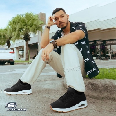 Homem sentado com sneakers pretos Skechers, calças claras e camisa preta com padrão branco CULTURE