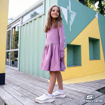 Menina com vestido rosa e sapatilhas Skechers junto a parede colorida e porta de vidro