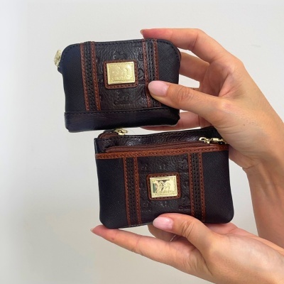 Duas carteiras pequenas pretas com detalhes em pele castanha e emblemas dourados, seguradas por mãos femininas.