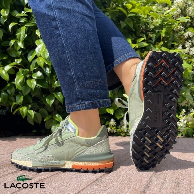 Sapatilhas Lacoste verdes com sola preta e detalhes laranja, usadas com calças de ganga em pavimento exterior