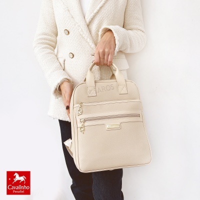 Mala bege de mão em pele sintética com bolso frontal e fechos dourados, segurada por pessoa com casaco branco felpudo