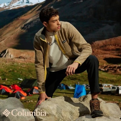 Homem com casaco bege Columbia em paisagem montanhosa com tendas e neve ao fundo