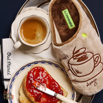 Chinelo de interior bege com desenho de chávena de café ao lado de chávena de café, torradas com compota e guardanapo branco num tabuleiro metálico.
