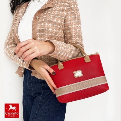 Mala de mão vermelha com detalhes em bege segurada por mulher com camisola castanha e camisa branca.