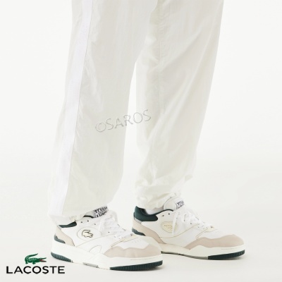 Ténis Lacoste branco, bege e verde escuro com calças brancas num fundo branco