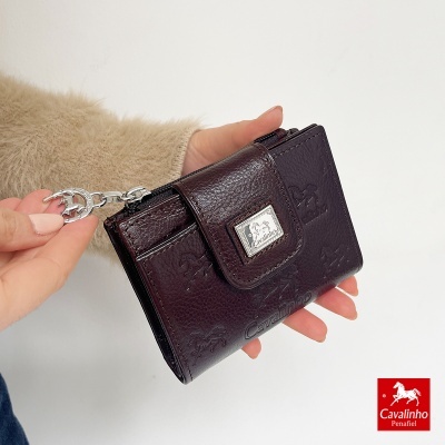 Carteira castanho escuro em couro com padrão gravado e fecho metálico, segurada por mão com roupa castanha clara