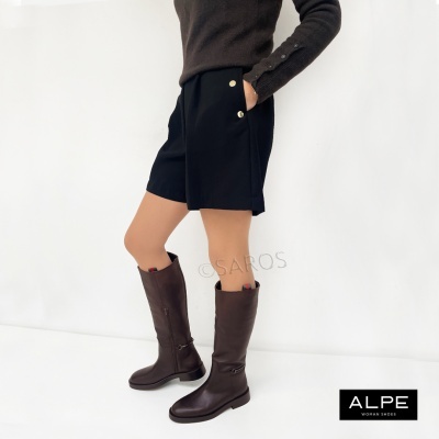 Pessoa com botas altas castanhas de couro e calções pretos numa parede branca