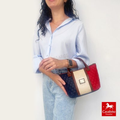 Mulher com blusa azul clara segurando mala de mão tricolor com detalhes em castanho.