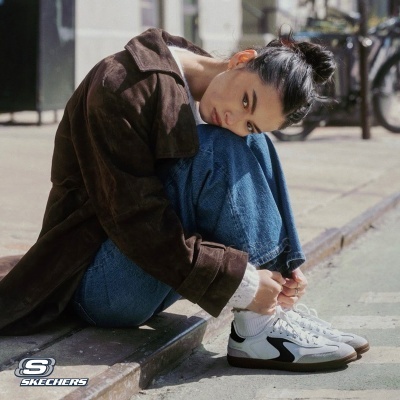 Mulher com casaco castanho, jeans azuis e sapatilhas brancas Skechers sentada na calçada