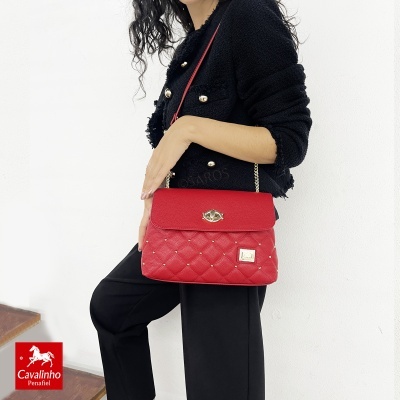 Mala de senhora vermelha de pele matelassê com fecho e alça dourados, vestida com casaco preto com botões dourados.