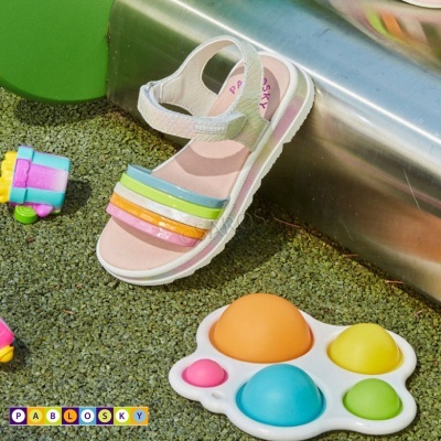 Sandália branca infantil com tiras coloridas e brinquedos coloridos num chão verde