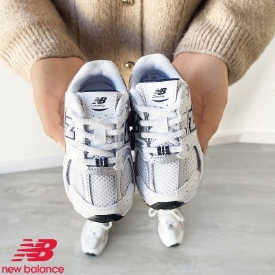 Ténis New Balance brancos com detalhes cinza e preto sendo segurados por duas mãos com casaco bege