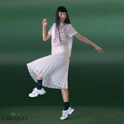 Mulher com vestido branco de malha transparente, meias pretas e sapatilhas brancas em fundo verde com logo Lacoste.