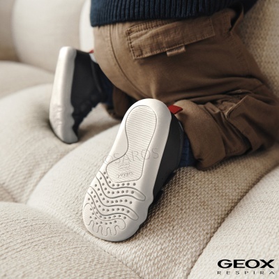 Sola branca antiderrapante de sapato infantil Geox em cima de sofá bege com criança vestindo calças castanhas.