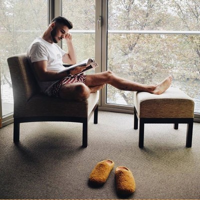 Homem sentado numa poltrona castanha, banco castanho, chinelos castanhos no chão junto a janela