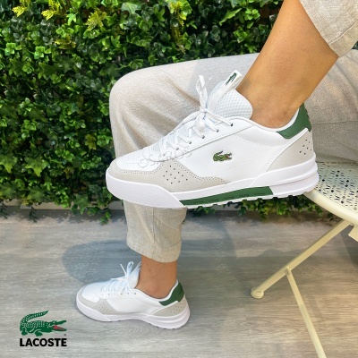 Tênis branco e verde Lacoste com logótipo de crocodilo ao lado de parede vegetal