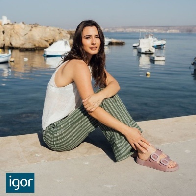 Mulher sentada à beira-mar com blusa branca, calças verdes às riscas e sandálias rosas