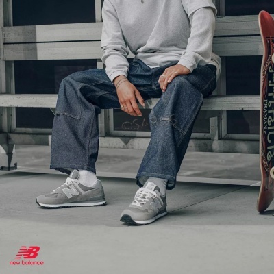 Sapatilhas New Balance cinzentas com detalhes brancos usadas por pessoa sentada num banco metálico.