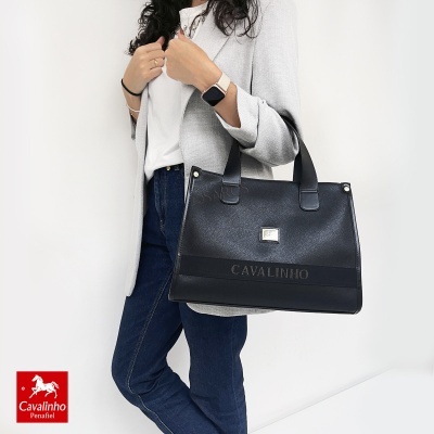 Mala preta Cavalinho em pele sintética com alças, logótipo metálico e texto CAVALINHO, usada por mulher com casaco cinza e jeans.