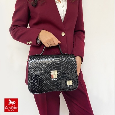 Mala de mão preta com padrão de pele de réptil e fecho dourado segurada por mulher com fato bordeaux