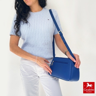 Mulher com camisola azul clara, calças brancas e mala de ombro azul escuro, fundo branco