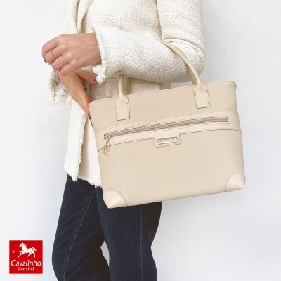 Mala de mão bege com bolso frontal e alças, segurada por pessoa com casaco bege claro e calças jeans