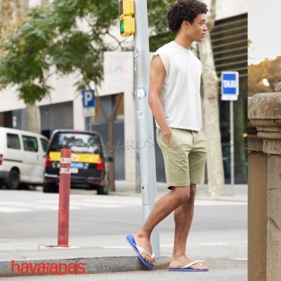 Homem jovem com t-shirt branca sem mangas, calções bege e chinelas azuis em rua urbana