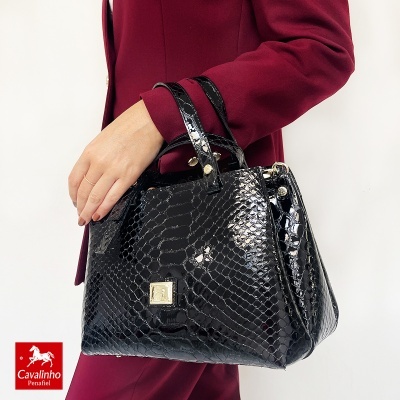 Mala de mão preta com textura de cobra brilhante e detalhe dourado, usada por pessoa com casaco bordô