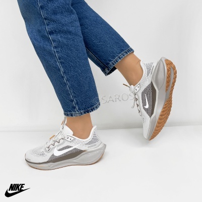 Ténis Nike brancos e cinzentos com sola castanha usados com jeans azuis em fundo branco