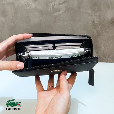 Carteira preta aberta em duas mãos mostrando interior branco e fecho com texto LACOSTE.