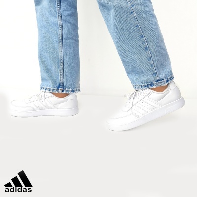 Ténis brancos Adidas com três riscas laterais usados com calças de ganga azul claro.