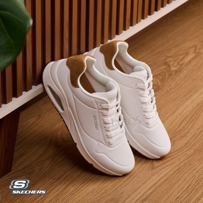 Sapatilhas brancas Skechers com detalhes castanhos junto a uma parede de madeira