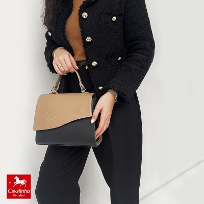 Mala de mão castanha e preta Cavalinho com detalhe de cavalo em relevo, segurada por pessoa com roupa preta e castanha