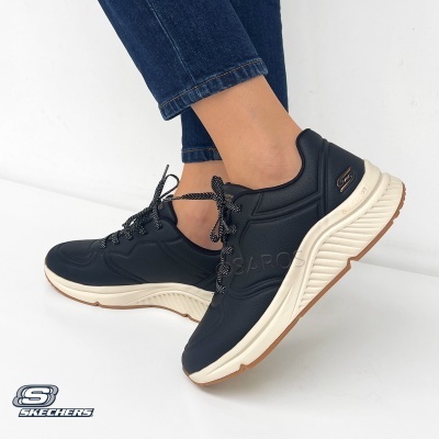 Ténis pretos com sola branca da marca Skechers em pés com jeans azuis