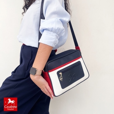 Mala Cavalinho branca, preta e vermelha com bolso frontal e alça preta, usada por mulher com camisa azul clara e calças escuras