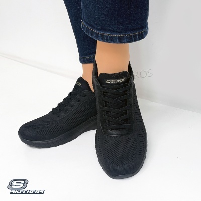 Ténis pretos Skechers com atacadores e sola preta usados por pessoa com jeans escuros.