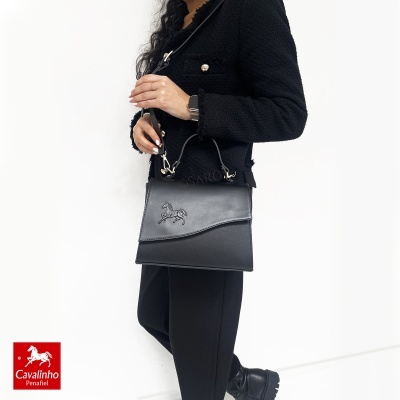 Mala preta de senhora em pele com símbolo de cavalo em relevo, usada por mulher vestindo casaco e calças pretas.