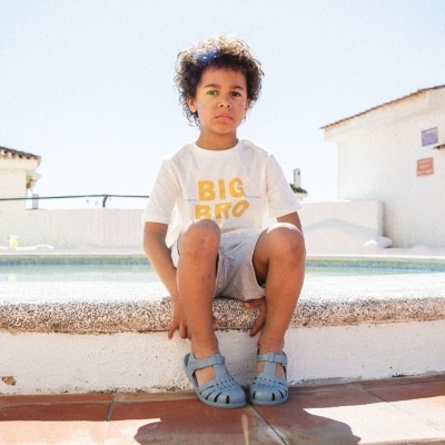 Menino sentado perto de piscina com sandálias azuis e t-shirt branca com texto amarelo