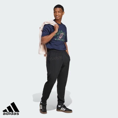 Homem com t-shirt azul, calças pretas, sapatilhas Adidas pretas e casaco bege sobre o ombro