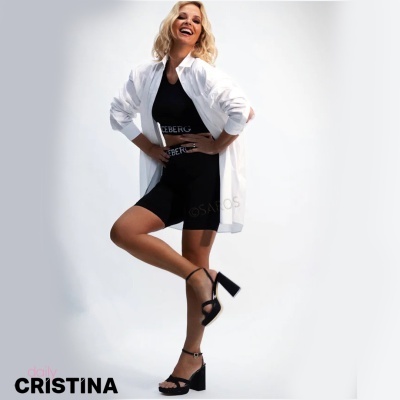 Mulher loira com vestido preto e camisa branca posando com sandálias de salto num fundo branco