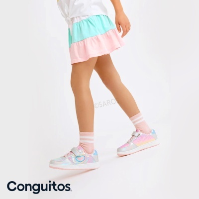 Ténis infantis multicoloridos com padrão brilhante e detalhe de coração nas laterais usados com meias cor-de-rosa listradas e saia rodada em rosa e azul claro