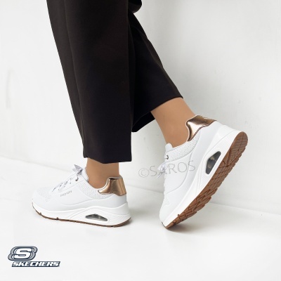 Sapatilhas brancas Skechers com detalhes dourados no calcanhar e sola castanha numa pessoa com calças pretas.