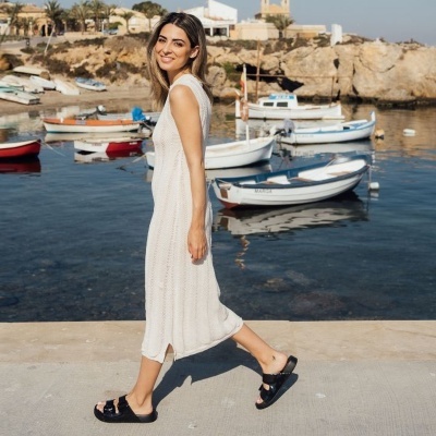 Mulher com vestido branco e sandálias pretas a caminhar junto ao mar com barcos atrás