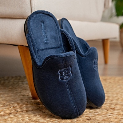 Chinelos de casa azuis escuros em tecido macio com bordados, em tapete de fibras naturais e sofá bege ao fundo