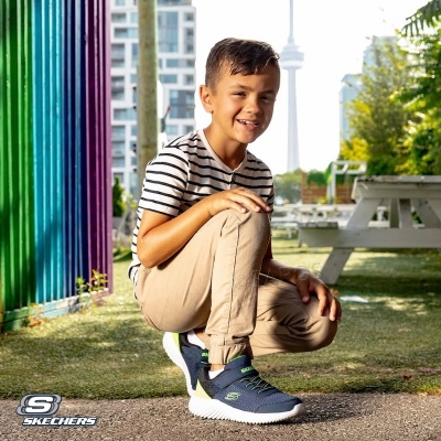 Menino com sapatilhas azuis Skechers, calças bege e t-shirt às riscas, em parque urbano com bancos e prédios ao fundo.