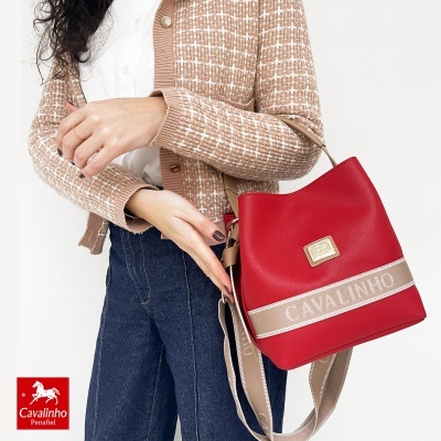 Mala vermelha com alça bege e faixa CAVALINHO usada por pessoa com casaco bege xadrez e calças jeans.