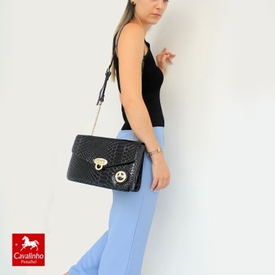 Bolsa preta textura crocodilo com fecho dourado, usada por mulher com top preto e calças azuis