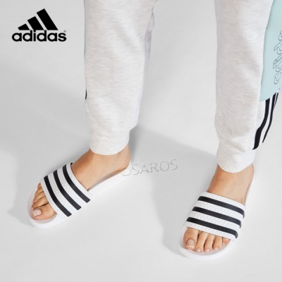 Chinelos brancos Adidas com riscas pretas usados com calças cinza claro.
