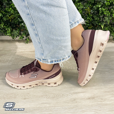 Ténis Skechers castanhos com sola branca usados com jeans claros, em piso de madeira e fundo com plantas.