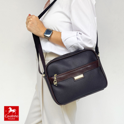 Bolsa quadrada preta com detalhes castanhos e fecho dourado exibida por pessoa com camisa branca